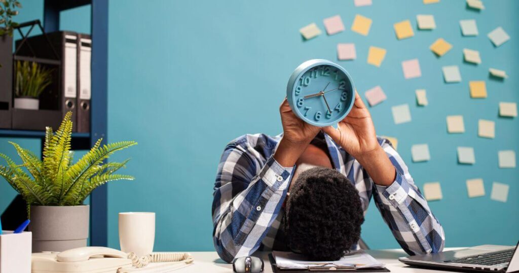 Empleado con la cabeza en el escritorio sosteniendo un reloj